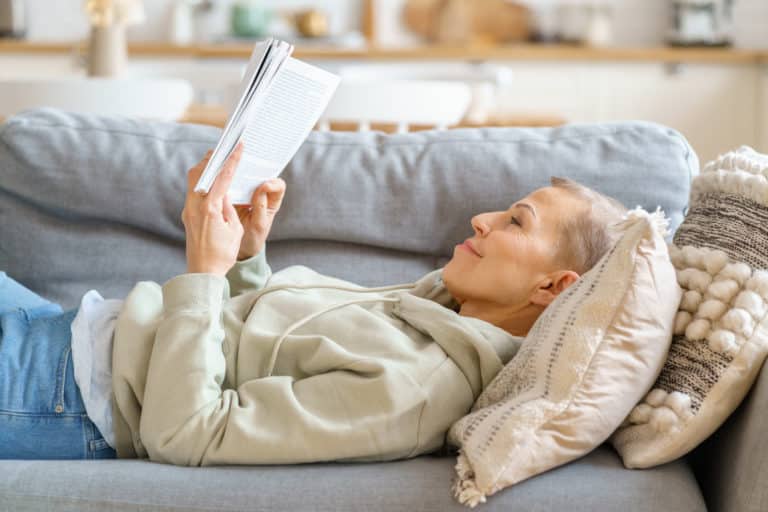 Cheerful middle aged woman reading interesting book and relaxing on sofa, spending leisure time alone at home. Selective focus on senior female